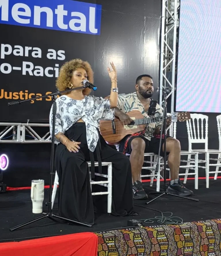 Cantora e violonista se apresentam no palco diante do painel do tema de 2025