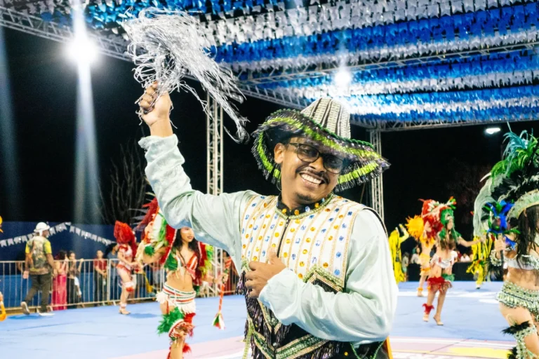 Brincante do Boi do Favela sorri com chapéu bordado e braço erguido durante apresentação noturna