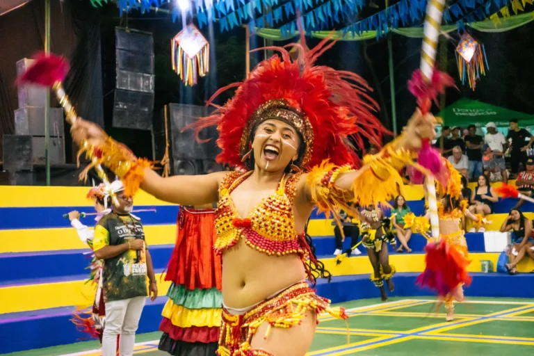 Dançarina do Boi do Favela abre os braços e sorri com cocar vermelho durante a apresentação