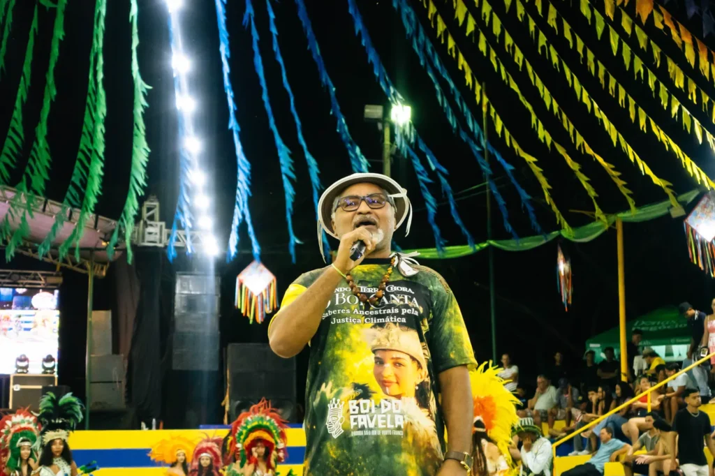 Narrador do Boi do Favela ao microfone, com a camiseta do projeto, conduzindo a história da apresentação