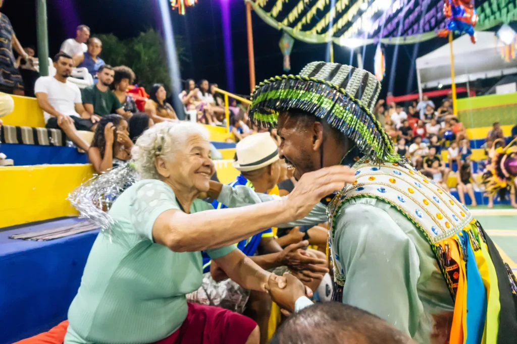 Senhora idosa dança e sorri ao lado de um brincante do Boi do Favela no meio do público