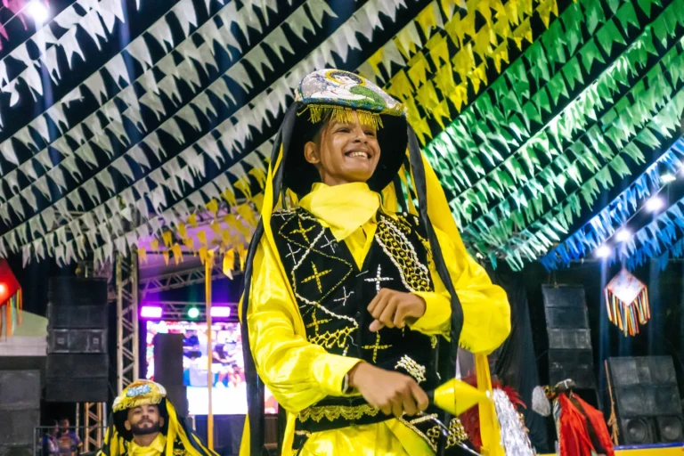 Brincante do Boi do Favela sorri de perto, com figurino amarelo bordado e chapéu
