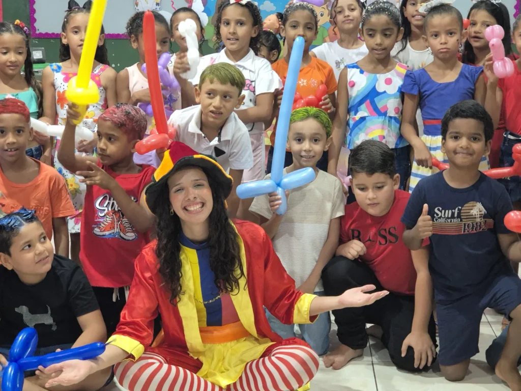 Recreadora fantasiada sorri cercada por crianças com balões coloridos em atividade da CIATDAL