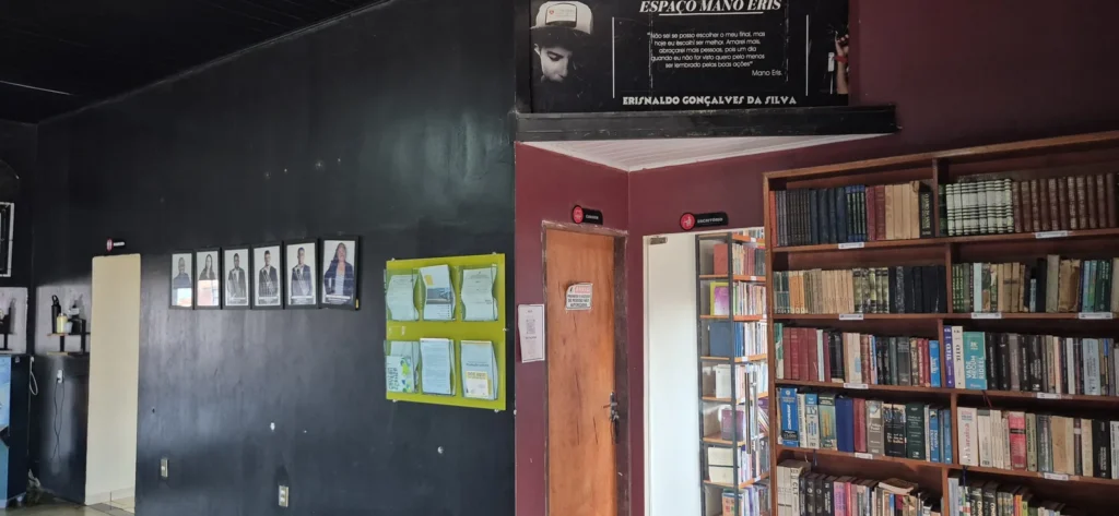 Painel do Espaço Mano Eris com o nome de Erisnaldo Gonçalves da Silva e estante de livros da biblioteca comunitária